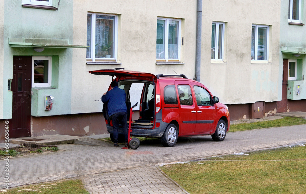 A car with an open trunk next to the residential building and a man unpacking goods