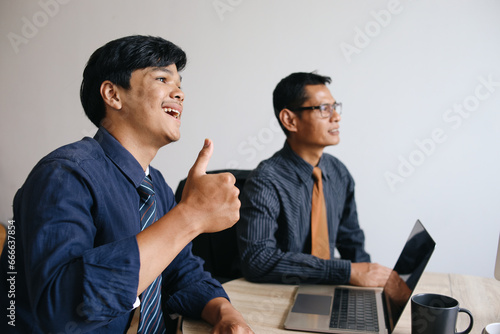 Young Asian smiling businessman showing thumbs up while sitting by senior male colleague in presentation meeting at the office
