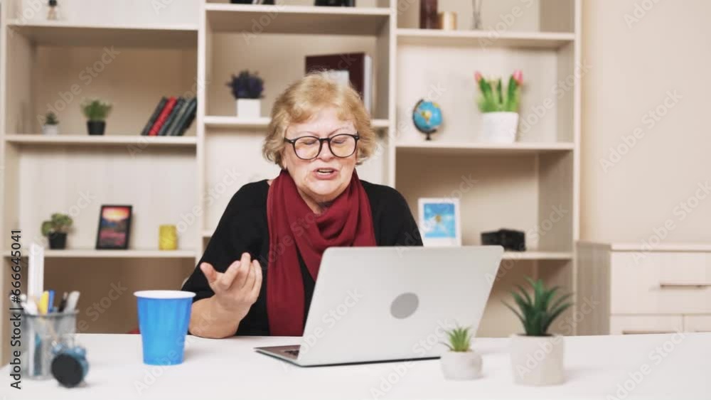 older woman in glasses is talking via video call while sitting at a table with a laptop older woman in glasses is talking via video call while sitting at a table with a laptop