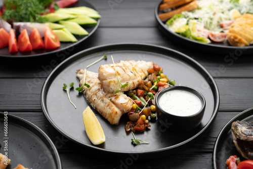 Grilled BBQ fish fillet with vegetables on black plate on dark wooden background.