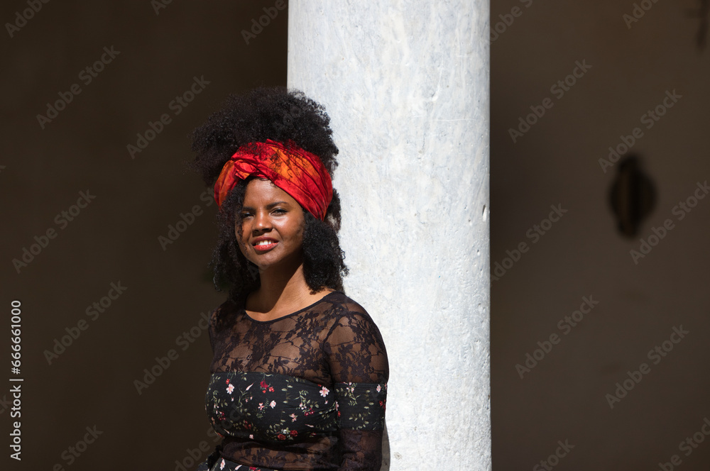 Φωτογραφία Portrait of a young, beautiful black woman with afro hair and black dress with flowers, wearing a red scarf in her hair