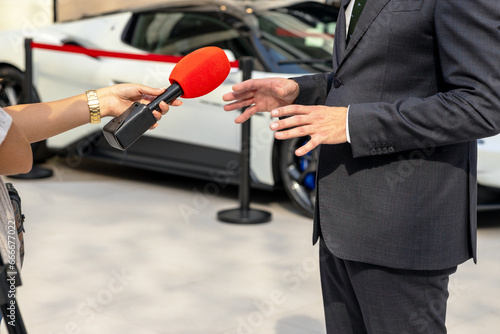 Media interview with car salesperson, sports car in the background