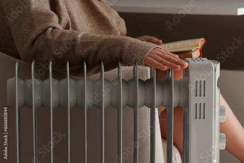 Woman in warm clothes is sitting next to the radiator. Woman is reading a book and warming her hands on an electric heater. It's cold at home. Energy crisis and high heating prices. 