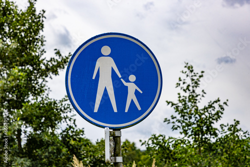 Pedestrian route or walking area sign against cloud covered sky and green foliage in background, blue metallic round border with drawing of an adult with a child walking in white, Netherlands