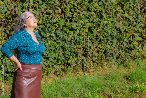 Elegant older adult woman standing in garden with her eyes closed and head tilted back sunbathing, brown skirt, blue blouse, long gray hair, hands on her waist, calm expression on her face