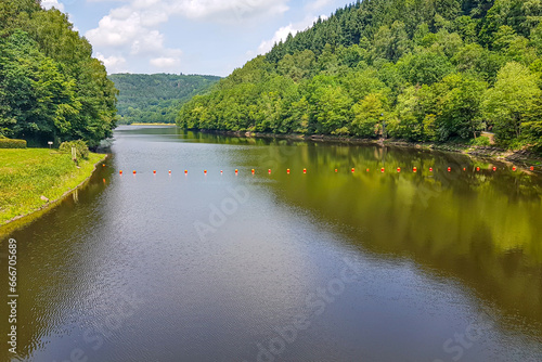 Landscape of Prum river with orange buoys on water surface in Stausee Bitburg reservoir  surrounded by abundant green leafy trees and mountains in background  sunny spring day in Germany