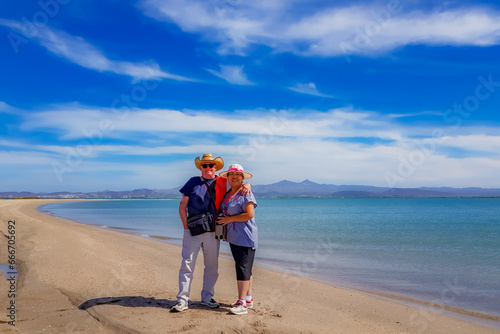 Senior adult tourist couple standing on beach sand with Gulf of Sea of Cortez and mountains in background, hat, sunglasses and casual clothes, sunny spring day in La Paz, Baja California Sur Mexico