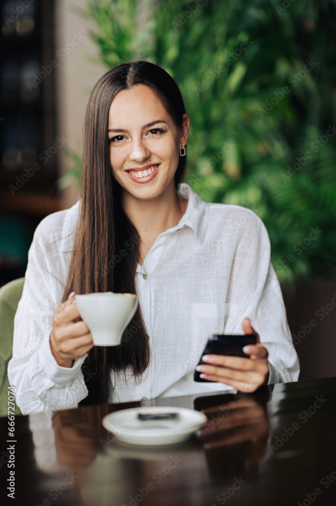 Cheerful brunette sitting in a coffee shop and enjoying her coffee while smiling at the camera. Cheerful brunette sitting in a coffee shop and enjoying her coffee while smiling at the camera.