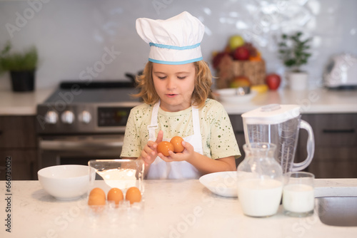 Child chef cook with eggs. Funny little kid chef cook wearing uniform cook cap and apron cooked food in the kitchen. Kids are preparing the dough, bake cookies in the kitchen.