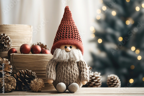 Close up of handcrafted christmas gnome knitted figurine on table against fir tree in living room, scandinavian, nordic dwarf with red hat. New Year accent decor in home interior. photo