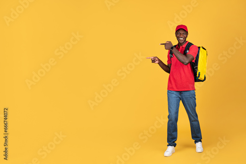 Glad millennial black guy courier in uniform with big backpack point finger at copy space