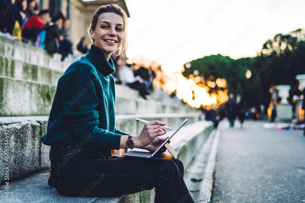 Fototapeta premium Portrait of cheerful hipster girl with electronic stylus and portable pc resting at city historic center
