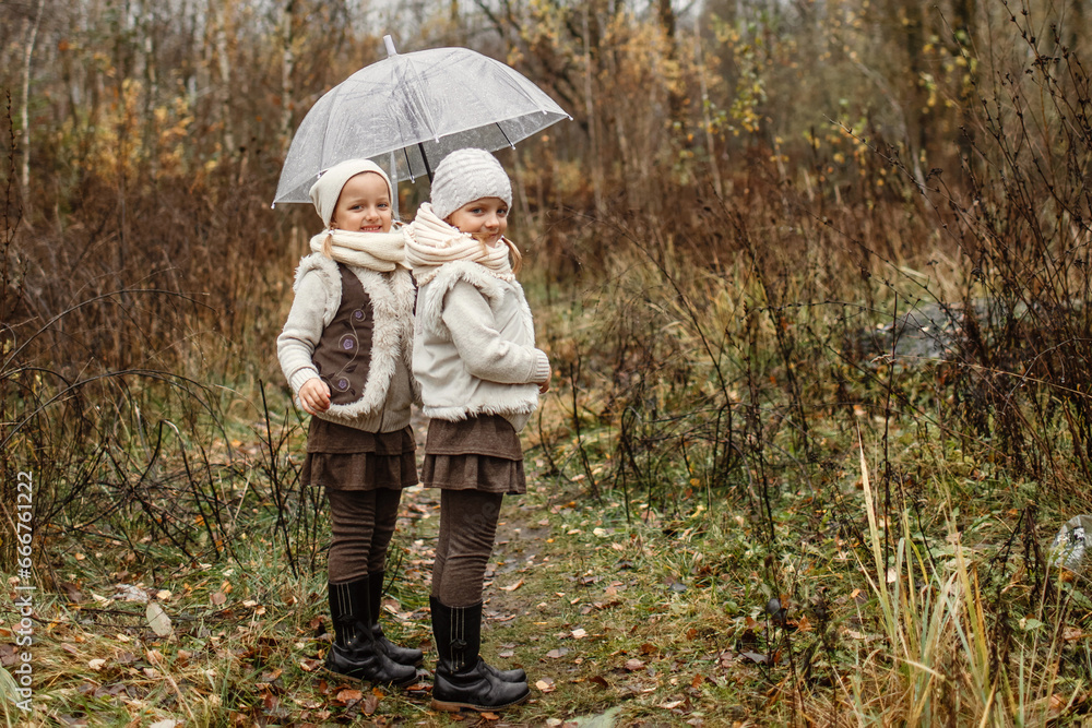 Fototapeta premium Portrait of young twin girls standing under an umbrella in the park, looking at the camera.