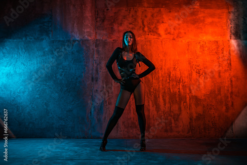 Young brunette woman dancer in black suit poses on wall background in red and blue light