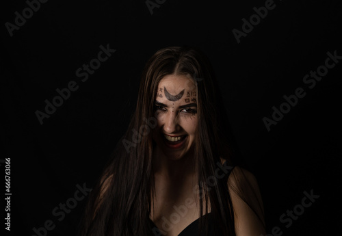 Evil laughing at the top of her voice, the witch looks into the camera on a black background in the twilight. On Halloween, a female witch grinning in a terrible grin looks at the camera.
