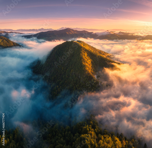 Aerial view of mountain peak in low clouds at sunrise in autumn. Top drone view of hills with red and orange trees in fog, colorful sky in fall. Slovenia. Nature. Mountain valley. Autumn forest. Alps