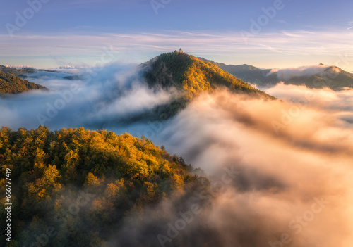 Aerial view of mountain peak in low clouds at sunrise in autumn. Top drone view of hills with red and orange trees in fog, colorful sky in fall. Slovenia. Nature. Mountain valley. Autumn forest. Alps