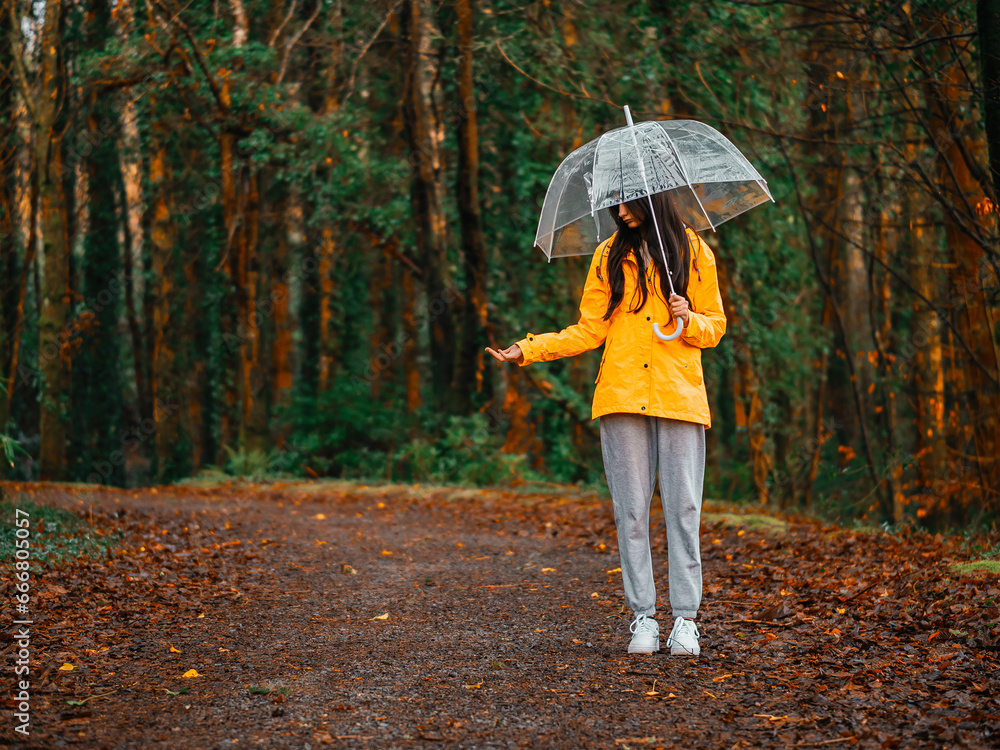Teenager girl in yellow jacket holding translucent umbrella and checking out rain while walking in a forest park. Outdoor activity and enjoy nature in any weather conditions.