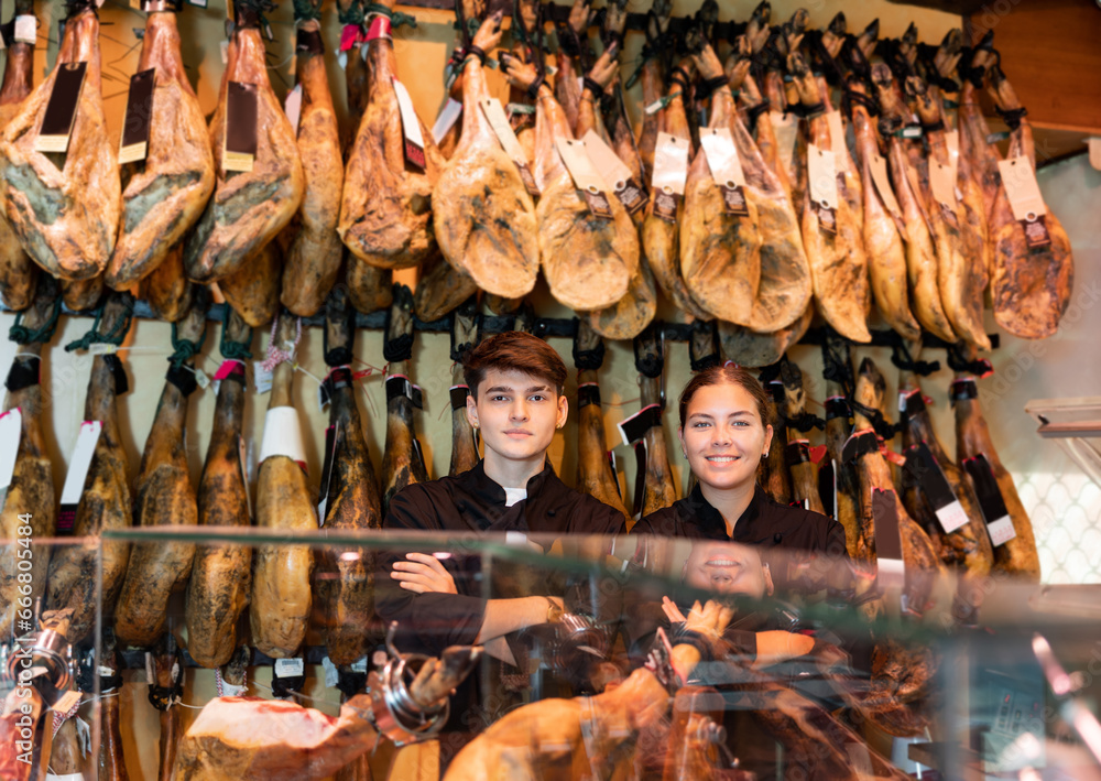 Two confident positive sellers of butcher shop, young girl and guy wearing black uniform, standing with crossed arms behind counter near rack with hanging whole legs of dry-cured Iberian jamon