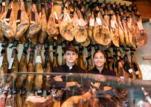 Two confident positive sellers of butcher shop, young girl and guy wearing black uniform, standing with crossed arms behind counter near rack with hanging whole legs of dry-cured Iberian jamon