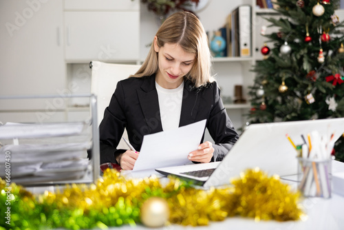 Smiling woman employee working in office with documents and laptop during Christmas time © JackF
