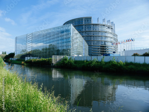 A sweeping view captures the Parliament building, gracefully bordered by a serene water canal, with all EU flags fluttering elegantly in the wind.