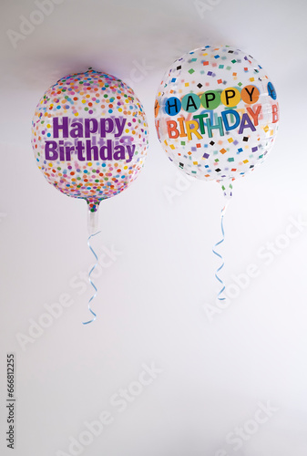 two helium balloons decorated with colored dots and a happy birthday message, with a hanging ribbon, on the roof of a white room, copy space, vertcial photo