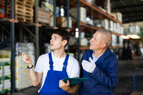 Elderly man and young guy warehouse workers in uniform discuss documents