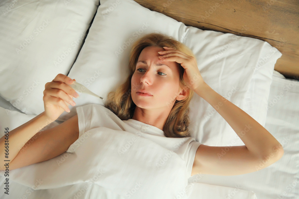 Sick woman measuring checking body temperature looking at thermometer while lying on bed at home Sick woman measuring checking body temperature looking at thermometer while lying on bed at home
