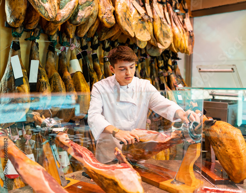 Smiling young guy seller in white uniform working at counter in butcher store, cutting slices from whole leg of dry-cured Iberian jamon on jamonera photo