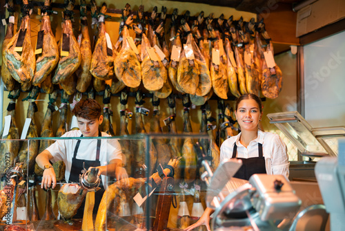 Young woman and man in uniform cutting ham with special knife. Woman and man selling traditional Spanish dish jamon.
