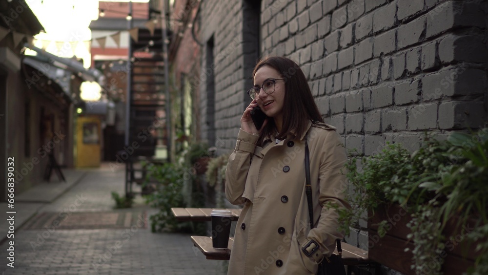 A young woman talks on the phone while standing against a wall on a narrow street. A girl with glasses and a coat speaks on the phone.