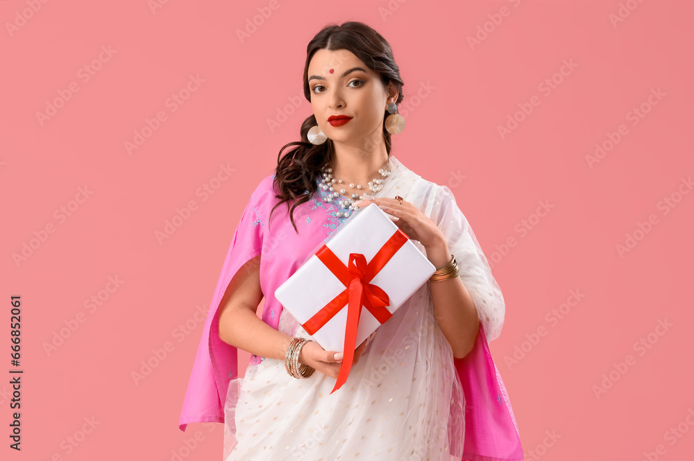 Fototapeta premium Beautiful young Indian woman in sari with gift box on pink background