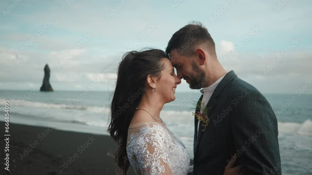 Loving smiling bride and groom with closed eyes standing face to face on beach with black sand. Blue sky, sharp rocks and hissing sea in the background. High quality 4k footage Loving smiling bride and groom with closed eyes standing face to face on beach with black sand. Blue sky, sharp rocks and hissing sea in the background. High quality 4k footage