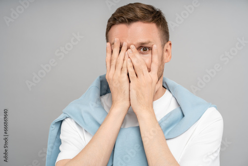 Shy modest playful guy hiding closing face with hands one eye looking at camera on isolated grey background. Portrait bashful timid coy man burns with shame feels awkward. Sincere emotions concept. photo