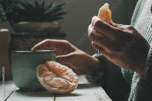 Close up of woman eating orange fruit and drinking herbal tea or coffee at home. healthy food and beverage lifestyle people indoor. Dark mood color green grey. Relaxation and enjoying relax inside