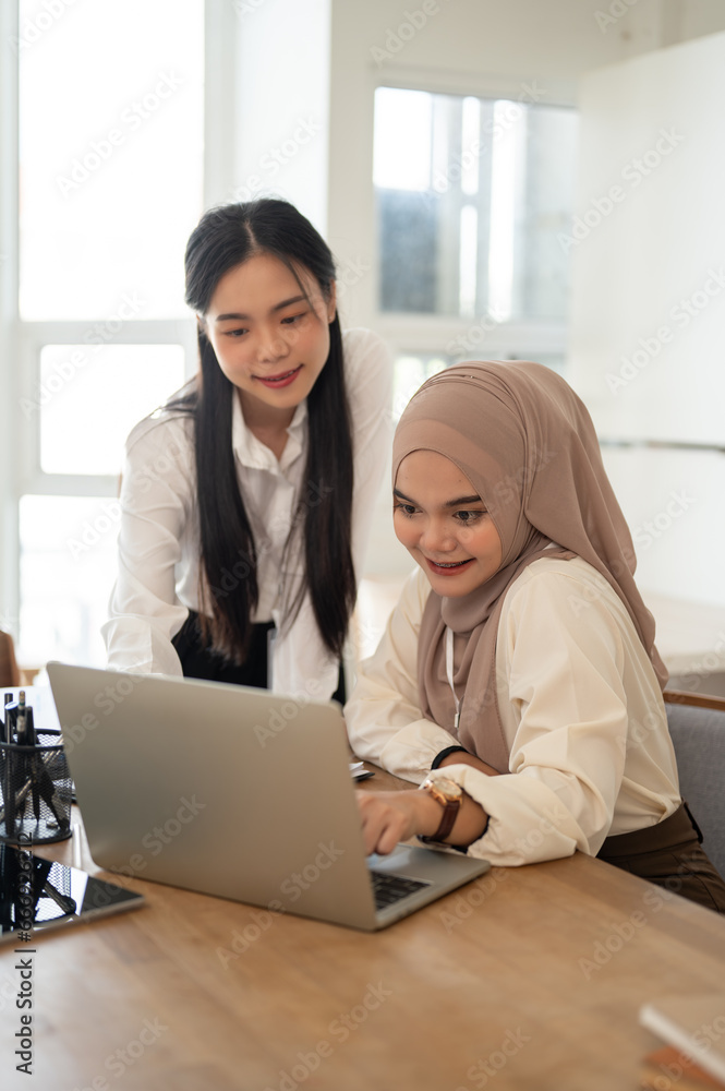 A beautiful Asian-Muslim businesswoman using her laptop while discussing work with her colleague. A beautiful Asian-Muslim businesswoman using her laptop while discussing work with her colleague.