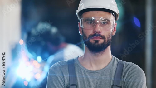 Portrait of a professional heavy industry engineer. Worker wearing protective uniform, goggles and helmet, smiling. In the background is a disoriented large industrial plant where welding sparks fly. photo