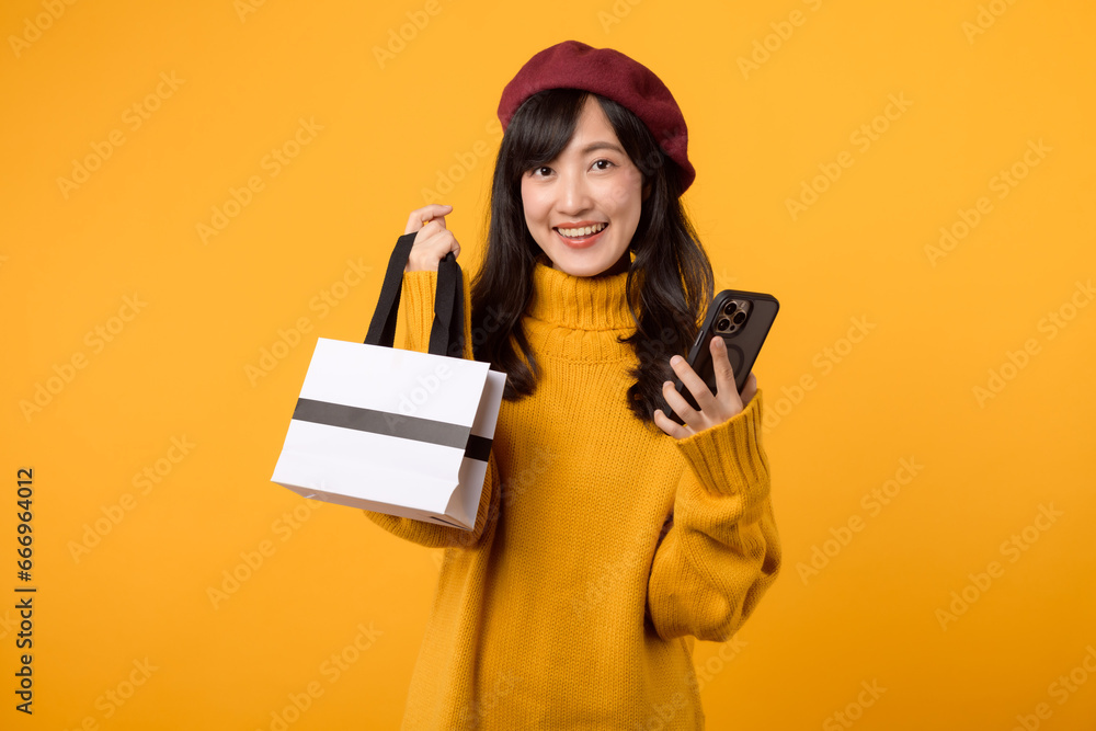 Fashion-forward young Asian woman in her 30s, holding a shopping paper bag and smartphone, celebrating her purchase while wearing a yellow sweater and red beret against a yellow background. Fashion-forward young Asian woman in her 30s, holding a shopping paper bag and smartphone, celebrating her purchase while wearing a yellow sweater and red beret against a yellow background.
