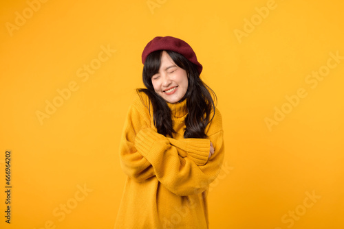 Shy young Asian woman in her 30s, arms crossed on her chest, with a charming smile, dressed in a yellow sweater and red beret against a yellow background.