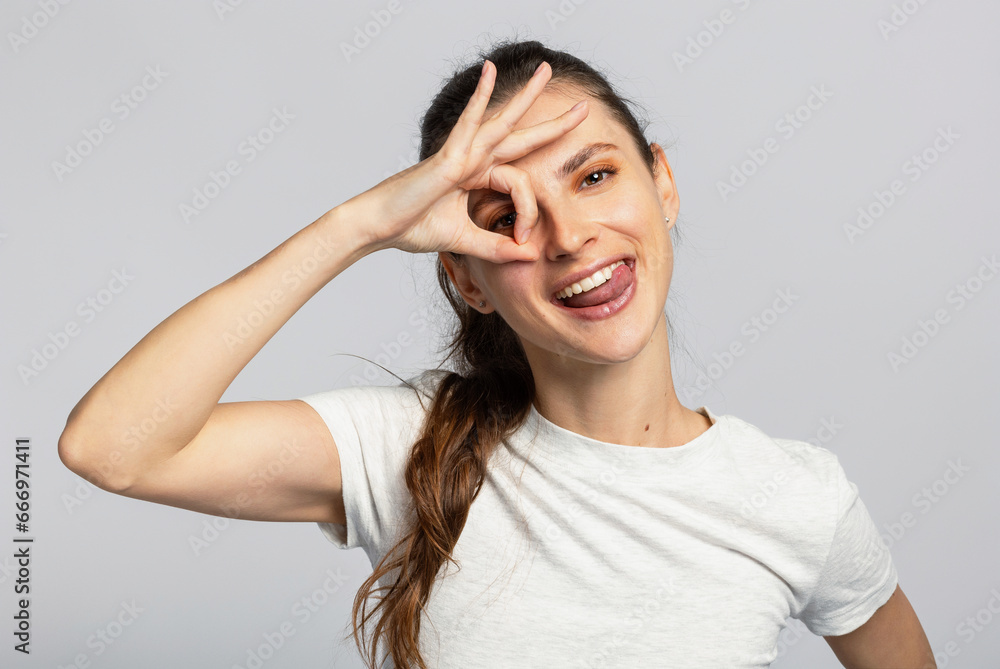 Excited young attractive woman showing okay gesture near her eye, on light grey studio background Excited young attractive woman showing okay gesture near her eye, on light grey studio background
