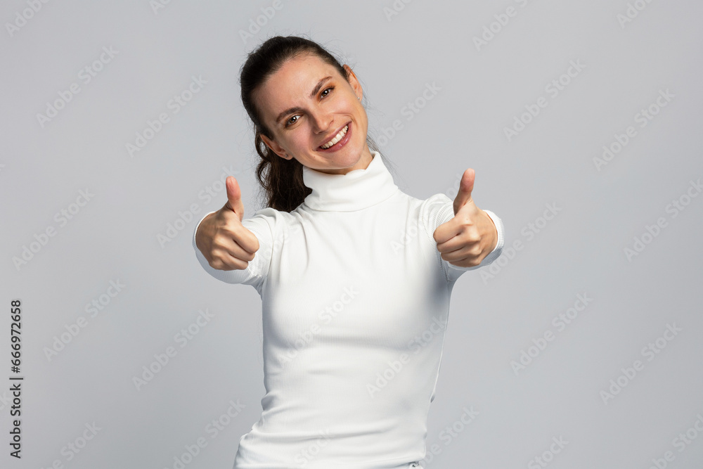 Joyful young woman make positive sign with her thumb up, positive mood, successful gesture, on light grey studio background Joyful young woman make positive sign with her thumb up, positive mood, successful gesture, on light grey studio background