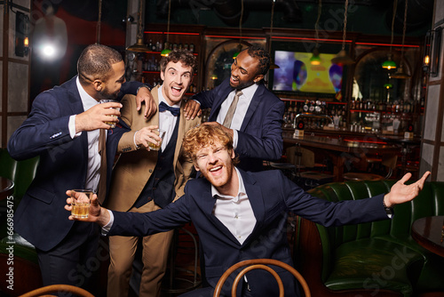 happy interracial friends in formal wear having bachelor party and holding whiskey in bar, diversity