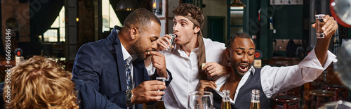 banner of funny and drunk multicultural men in formal wear drinking tequila in bar after work photo