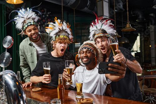 happy multicultural men in headwear with feathers taking selfie on smartphone during bachelor party