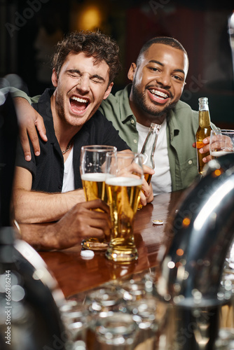 happy african american man hugging excited friend and holding glass of beer in bar, male friendship