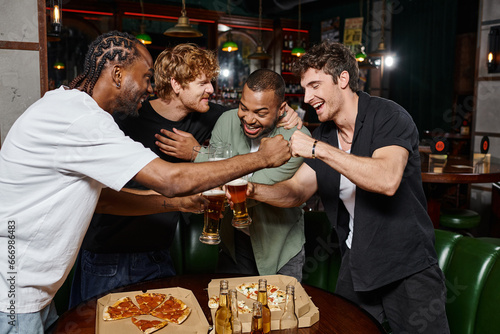 happy interracial male friends fist bumping while toasting glasses of beer in bar, bachelor party