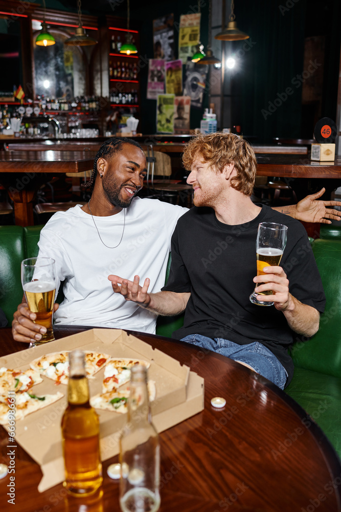 happy redhead man gesturing while chatting with african american friend over beer and pizza in bar