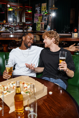 happy redhead man gesturing while chatting with african american friend over beer and pizza in bar