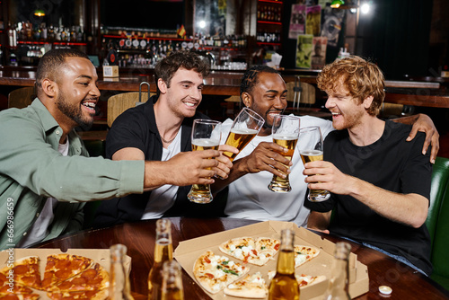 night out, joyful multiethnic men clinking glasses of beer near pizza in bar, male friendship